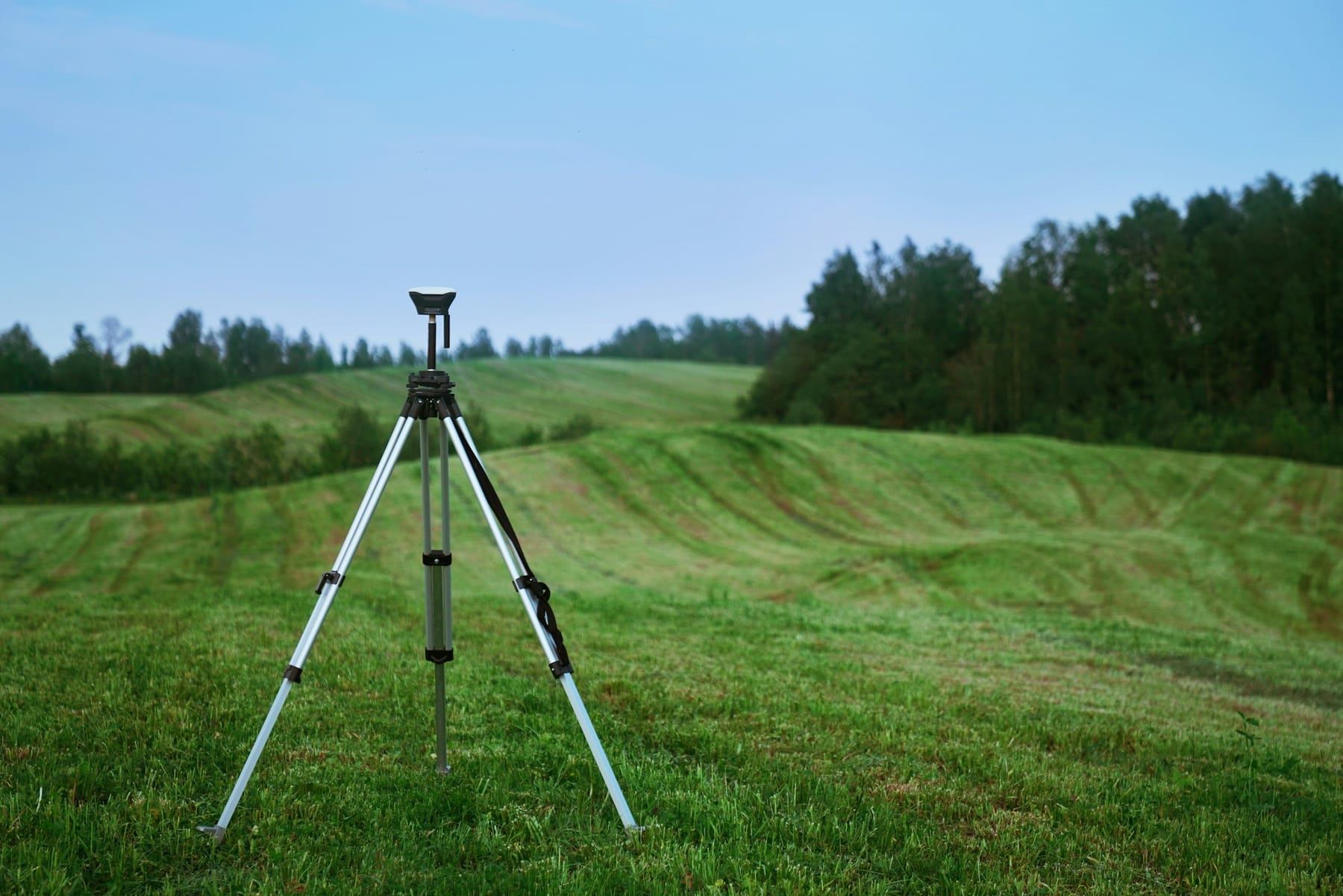 A survey instrument on a tripod standing alone in an empty grass field, with treeline behind and nobody in the frame.