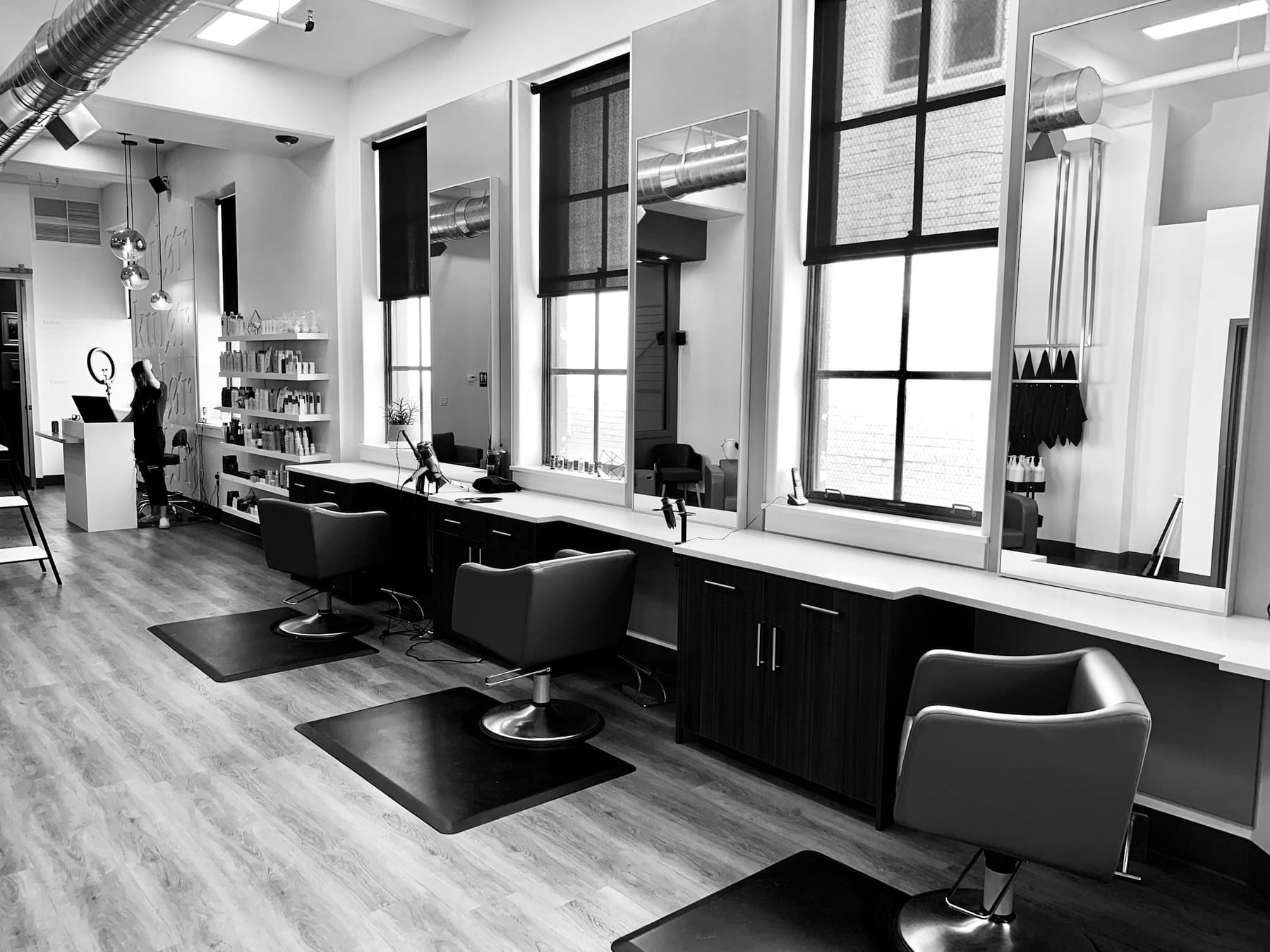 A salon with a row of chairs and mirrors along a wall of tall windows.