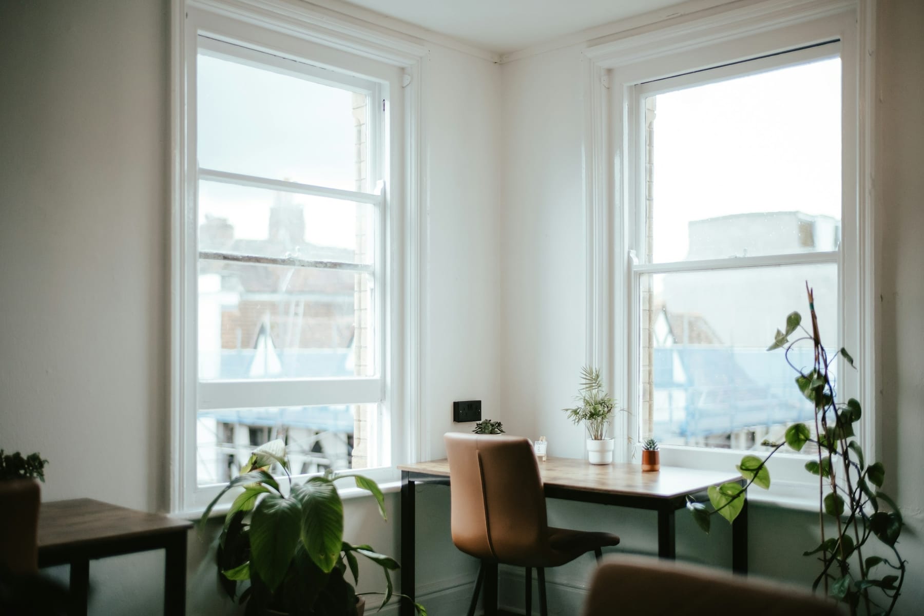 A single small desk and chair between two bright windows in a plain white room, with a plant on the sill.